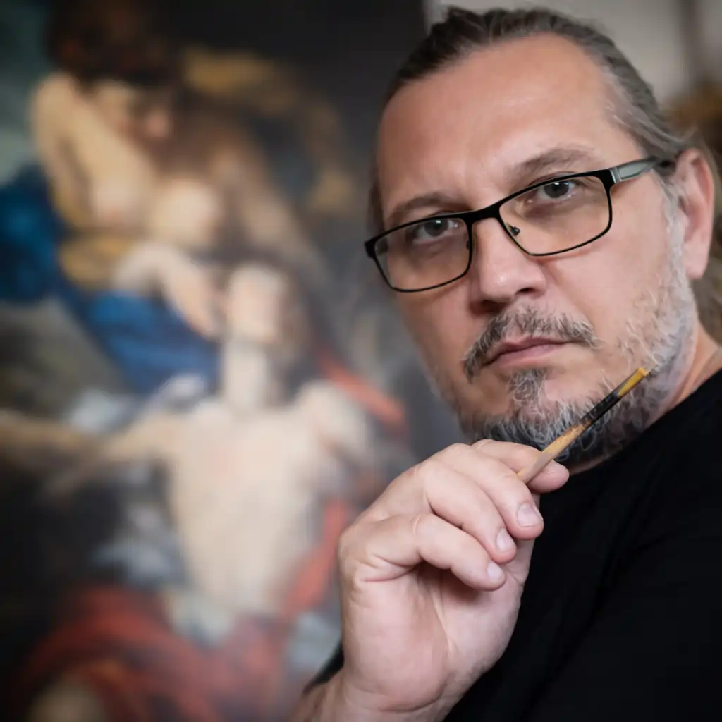 Artistul Călin Bogătean ținând pensula în atelier, cu pictură clasică în fundal, lucrări realizate la comandă Călin Bogătean pictând icoană pe lemn în atelier – icoane pe lemn pictate manual Contact – Călin Bogătean